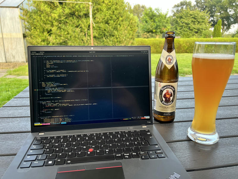 In the front a laptop on a table, a glas of beer. In the background a garden and some bushes.
