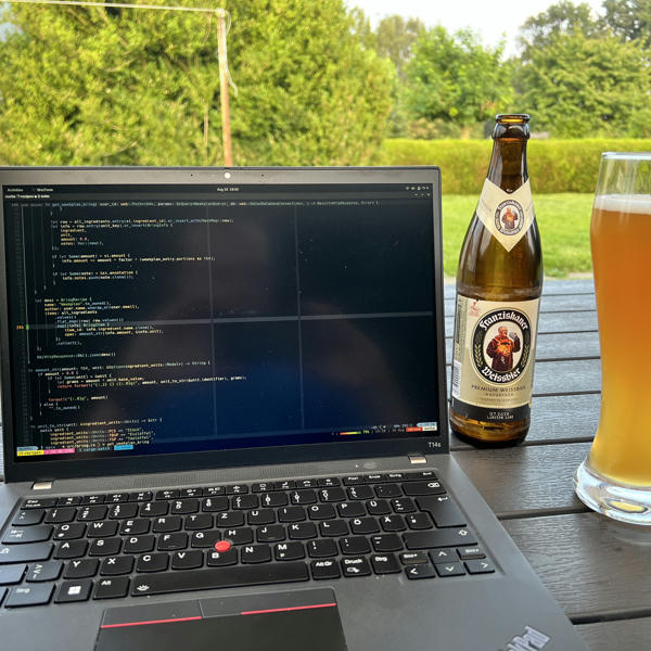 In the front a laptop on a table, a glas of beer. In the background a garden and some bushes.